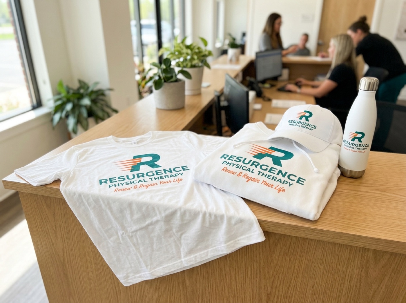 Resurgence Physical Therapy branded white apparel — t-shirt, hoodie, cap, and water bottle on a clinic reception desk